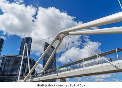 A low angle view of a white modern pedestrian bridge and sleek skyscrapers against a vibrant blue sky with fluffy clouds.