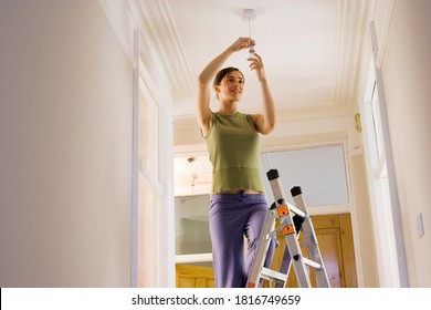 Low angle view of a woman doing DIY at home standing on ladder and attaching light bulb to the ceiling fixture. - Powered by Shutterstock