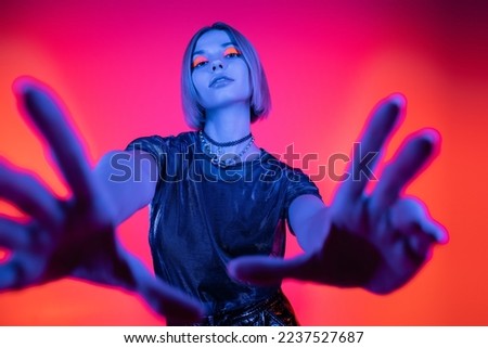 low angle view of woman with glowing makeup and outstretched hands in blue neon light on coral and pink background
