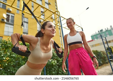 Low angle view of woman helping girlfriend doing exercise with trx suspension straps for back on sports ground. Caucasian athletic girls wearing sportswear. Healthy lifestyle. Willpower. Friendship