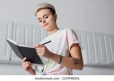 Low Angle View Of Woman With Trendy Hairstyle Writing In Notebook In Kitchen