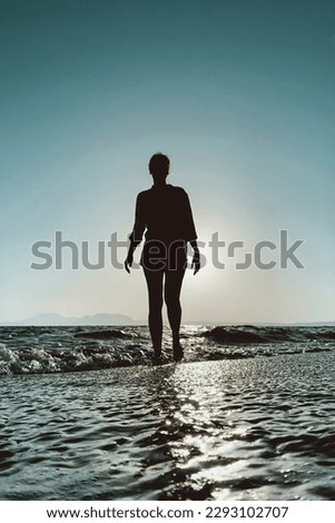 Low angle view of woman walking in backlit on a beach at sunset infront of the sun against blue sky