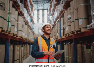 Low angle view of young african man wearing reflective jacket holding digital tablet standing in factory warehouse smiling