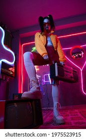 Low Angle View Of Young Asian Woman In Sunglasses And Orange Jacket Posing With Retro Boombox Near Tv