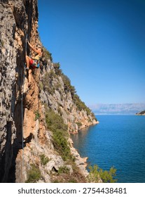 LOW ANGLE VIEW: Young athletic woman rock climbing on a majestic cliff overlooking the Adriatic Sea. Vastness and diversity of scenic Croatian coastline and the thrill of adrenaline sport climbing.