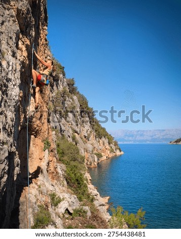 LOW ANGLE VIEW: Young athletic woman rock climbing on a majestic cliff overlooking the Adriatic Sea. Vastness and diversity of scenic Croatian coastline and the thrill of adrenaline sport climbing.