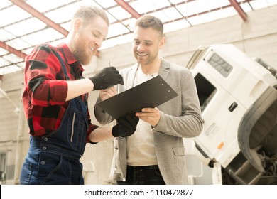 Low angle view of young bearded car mechanic showing inspection checklist to male client in service garage and smiling cheerfully