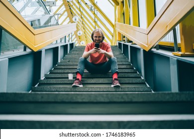 Low angle view of young bristled man looking at phone screen and smiling while sitting at stairs of enclosed pedestrian bridge - Powered by Shutterstock