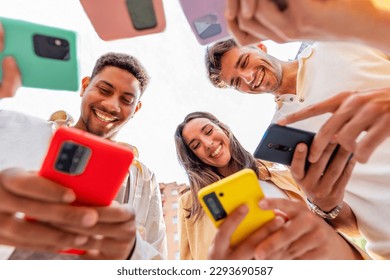 Low angle view of young group of millennial people in circle using smart mobile phones together outdoors. Joyful student friends looking at smartphone screen enjoying social media content - Powered by Shutterstock