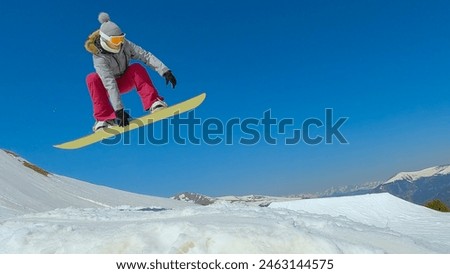 LOW ANGLE VIEW: Young lady on snowboard doing grab trick while jumping on kicker. Female snowboarder performing stylish jump trick in snow park at mountain ski area. Sunny winter day for snowboarding.