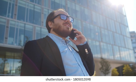 Low Angle View Of Young Obese Businessman Talking On Smartphone In City Outdoors. Chubby Positive Employee In Glasses Having Phone Conversation Standing Over Business Center Background