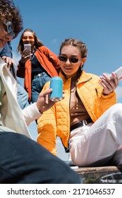 Low Angle View Of Young And Trendy Friends With Gadgets And Soda Cans Against Blue Sky