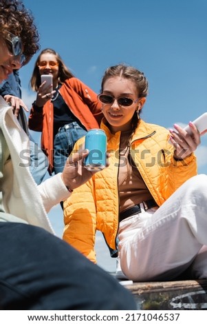 low angle view of young and trendy friends with gadgets and soda cans against blue sky