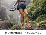 Low angle view of young woman walking down the mountain barefoot. Female hiker walking barefoot on rock with waterfall in background.