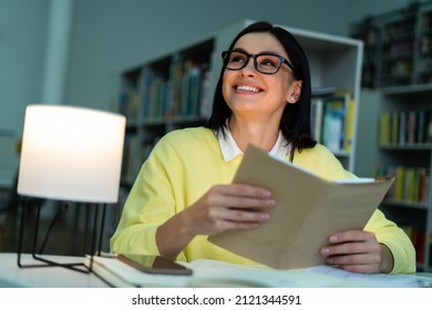 Low Angle View Of The Young Woman Reading Book At The Library And Smiling Toothy. Brunette Lady Studying At The High School. Stock Photo 