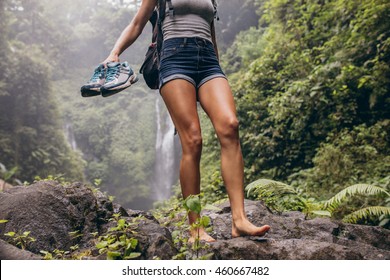 Low angle view of young woman walking down the mountain barefoot. Female hiker walking barefoot on rock with waterfall in background.