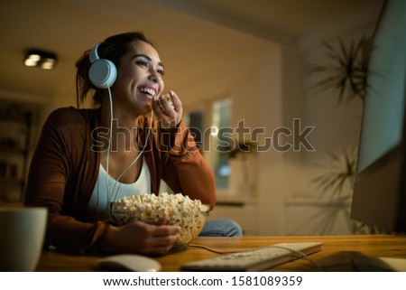 Low angle view of young woman having fun while eating popcorn and watching movie online on desktop PC at home. 