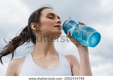 low angle view of young woman in crop top drinking water against blue sky with clouds