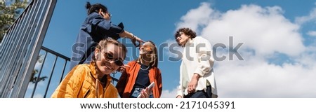 low angle view of young woman smiling at camera near interracial friends against cloudy sky, banner