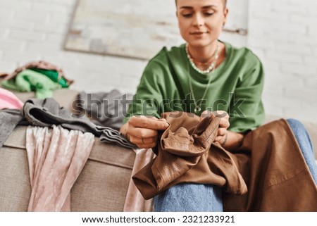 low angle view of young woman holding stylish leather pants while sitting on couch in living room and sorting clothes at home,  blurred background, sustainable living and mindful consumerism concept