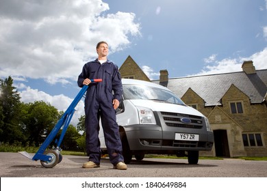 A Low Angle Wide Shot Of A Man In Coveralls Posing And Holding A Hand Truck On A Side With A White Work Van In Background.