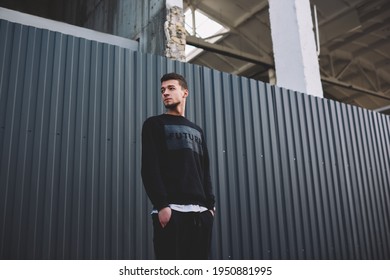 Low angle of young ethnic slender bearded male in trendy clothes keeping hands on pockets and looking away beside metal fence on street