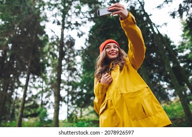 Low Angle Of Young Happy Female In Bright Yellow Rain Coat And Red Hat Taking Selfie Using Mobile Phone In Park