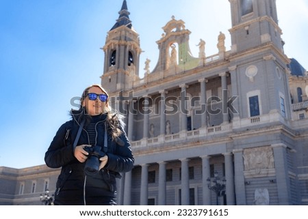Low angle of young Hispanic woman tourist in warm clothes and sunglasses standing with professional photo camera while looking away in sunlight near Cathedral La Almudena in Madrid, Spain