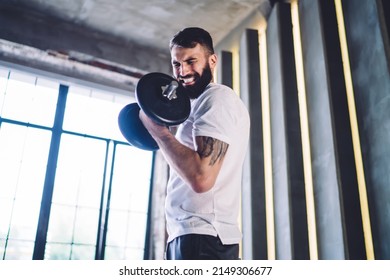 Low angle young man in sportswear with effort lifting heavy dumbbell for developing biceps muscles during training in morning in modern gym