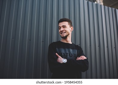 Low angle of young smiling bearded male in trendy clothes standing beside high metal fence on street and looking away