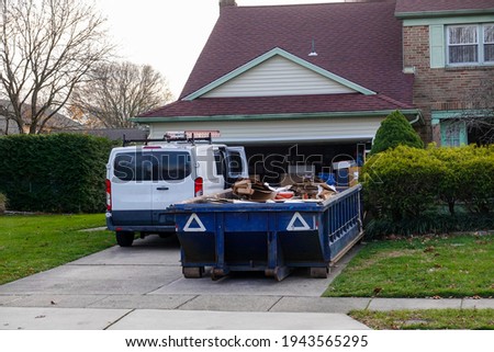 Low blue dumpster full of cardboard and construction debris in front of a full garage and next to a white worker's van
