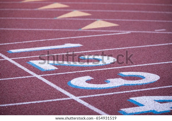 Low closeup on track and field crushed red rubber\
running surface divided into racing straight and arched lanes\
labeled by painted numbers with starting line marked by white\
stripe at athletic stadium
