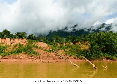 Low clouds hover over a muddy riverbank along the Nam Ou River near Muang Ngoi in northern Laos. Eroded cliffs, fallen tree trunks, and dense green vegetation create a textured landscape beneath a dramatic sky.