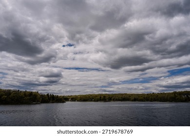 Low gray clouds stretch over a calm lake with a distant tree-lined shoreline and scattered houses. Gentle ripples move across the dark water