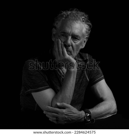 Low key black and white portrait of an attractive older Caucasian man with grey hair and beard. Turned to the right with a serious or pensive expression, hand over mouth, arms visible. 