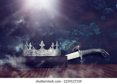 low key image of beautiful queen or king crown over antique book next to sword. fantasy medieval period. Selective focus