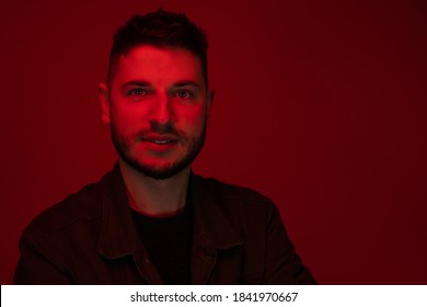 Low key portrait of bearded chubby young man looking at camera dressed casually in red light on dark red backdrop. UV ray skin damage