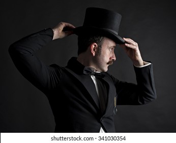 Low key side portrait of Victorian gentleman adjusting his top hat. He has a curled mustache and a gold monocle hanging from his breast pocket. 