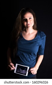 Low Key Studio Portrait Of A Happy Young Woman, In The Early Stages Of Pregnancy, Holding A Copy Of Her Scan In Front Of Her Body. All Identifying Marked Removed From Scan.