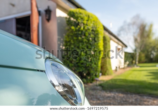 Low level view of a cars new style\
headlight. Seen parked in a private driveway, facing part of a\
large garden. A garage door can be seen opened on the\
left.