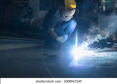 Low light image of Welder or craftsman in a welding mask and welders leathers, a metal product is welded with a welding machine in the garage,sparks fly to the sides.Factory concept.