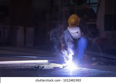 Low light image of Welder or craftsman in a welding mask and welders leathers, a metal product is welded with a welding machine in the garage,sparks fly to the sides. Setup studio shooting.