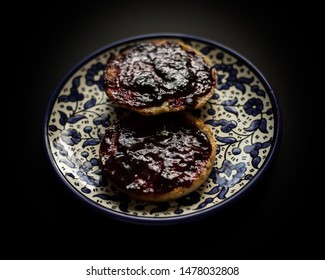 Low Light Photographed English Muffin Breakfast Topped With Blackberry Jam On A Blue China Plate.