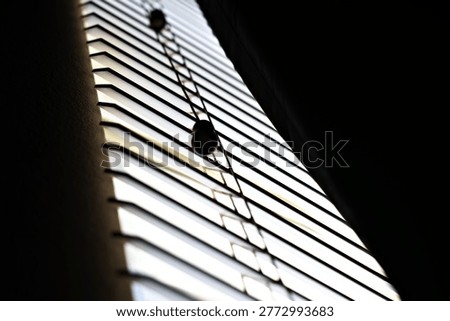 Low light through window blinds casts narrow shadows across a quiet interior, reflecting nightshift fatigue, isolation, and a subdued depressive mood.