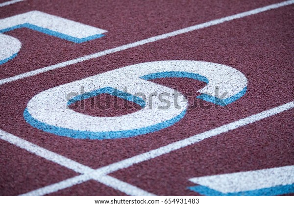 Low macro close up on track and field crushed red\
rubber running surface divided into racing lanes labeled by painted\
number six with starting line marked by white stripe at athletic\
sport stadium