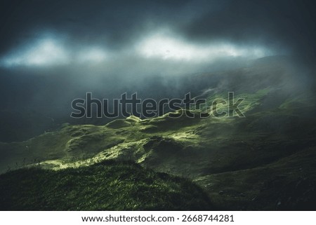 Low mist and storm clouds drift across the hills of Dovedale Valley, Lake District, with sunlight breaking through in a moody scene.