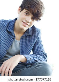 Low Perspective Portrait View Of An Attractive Teenager Boy Sitting Down With His Legs Crossed In A Park, Relaxing During A Sunny Day And Smiling At The Camera. Outdoors Lifestyle.