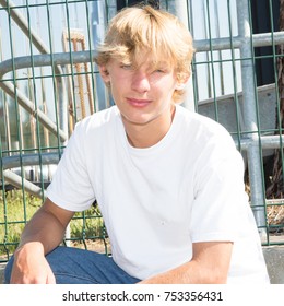 Low Perspective Portrait View Of An Attractive Teenager Boy Sitting Down With His Legs Crossed In A Park