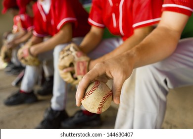 Low section of baseball team mates sitting in dugout with player holding a ball in foreground - Powered by Shutterstock