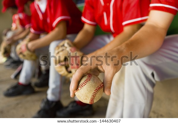 Low section of baseball team\
mates sitting in dugout with player holding a ball in\
foreground